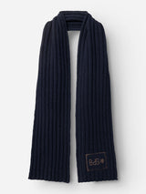 The BGcomforto Knit Scarf - Dark Navy by Blue de Gênes is a long, chunky knit scarf in a dark blue shade, featuring a small brown patch with stitched lettering near one end, displayed against a plain white background.