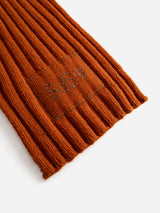 A close-up of the BGcomforto Knit Scarf - Ginger Bread Orange by Blue de Gênes, highlighting its ribbed wool texture, visible stitching, and a subtle embroidered patch with small symbols, showcasing the cozy chunky knit design.