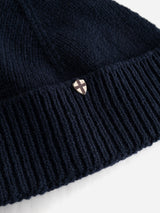 A close-up of the BGboshi Beanie in Dark Navy by Blue de Gênes, featuring a ribbed turn-up and a small, shield-shaped metal emblem with a cross design attached to the folded brim.
