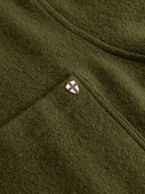 A close-up of the BGhew Wool Overshirt in Winter Moss by Blue de Gênes, showing its olive green, soft woolen texture and visible stitching, accented by a small silver shield-shaped pin featuring a white X design.