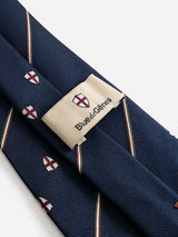 The BGshield Tie - Dark Navy by Blue de Gênes is a navy blue silk tie adorned with small red and white shield patterns, and features a beige label reading “Blue de Gênes” with the brand’s logo displayed above the text.