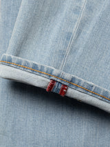 A close-up of BGvinci Trueblue Jeans by Blue de Gênes in mid blue denim, featuring a folded cuff with an orange seam and a red and blue striped fabric tag on the inner seam.