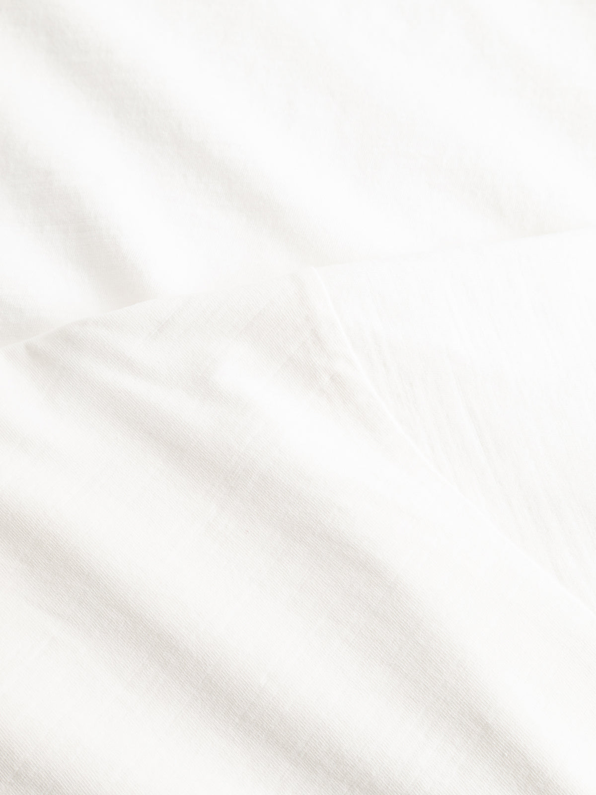 A close-up of smooth, white slub cotton fabric with subtle folds and soft shadows highlights the minimalist, clean look of Blue de Gênes' BGoscar T-Shirt - Snow Fall.
