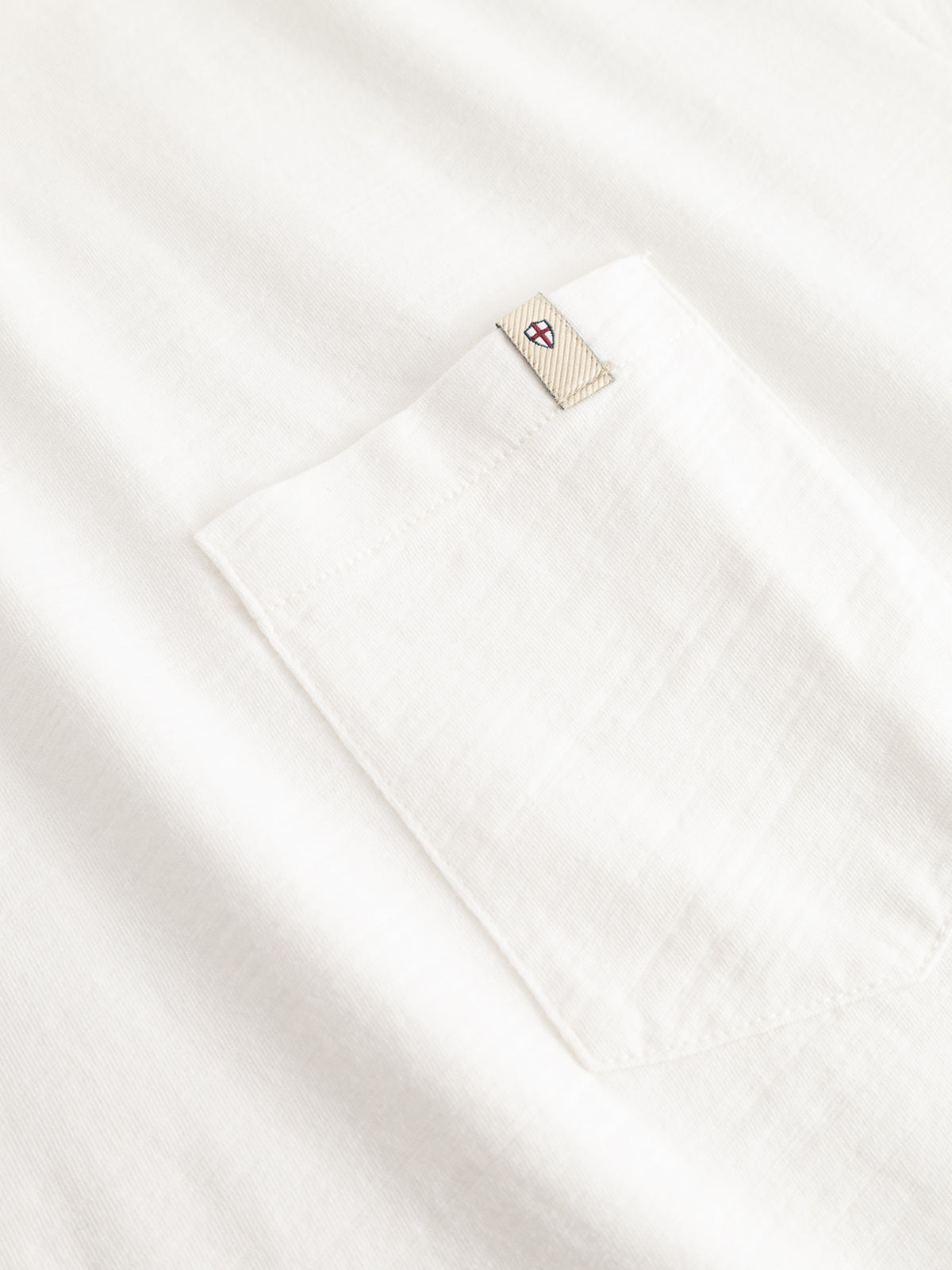 Close-up of the BGoscar T-Shirt - Snow Fall by Blue de Gênes, a white slub cotton tee with a chest pocket featuring a beige tag at the top and a small embroidered blue, red, and white emblem.