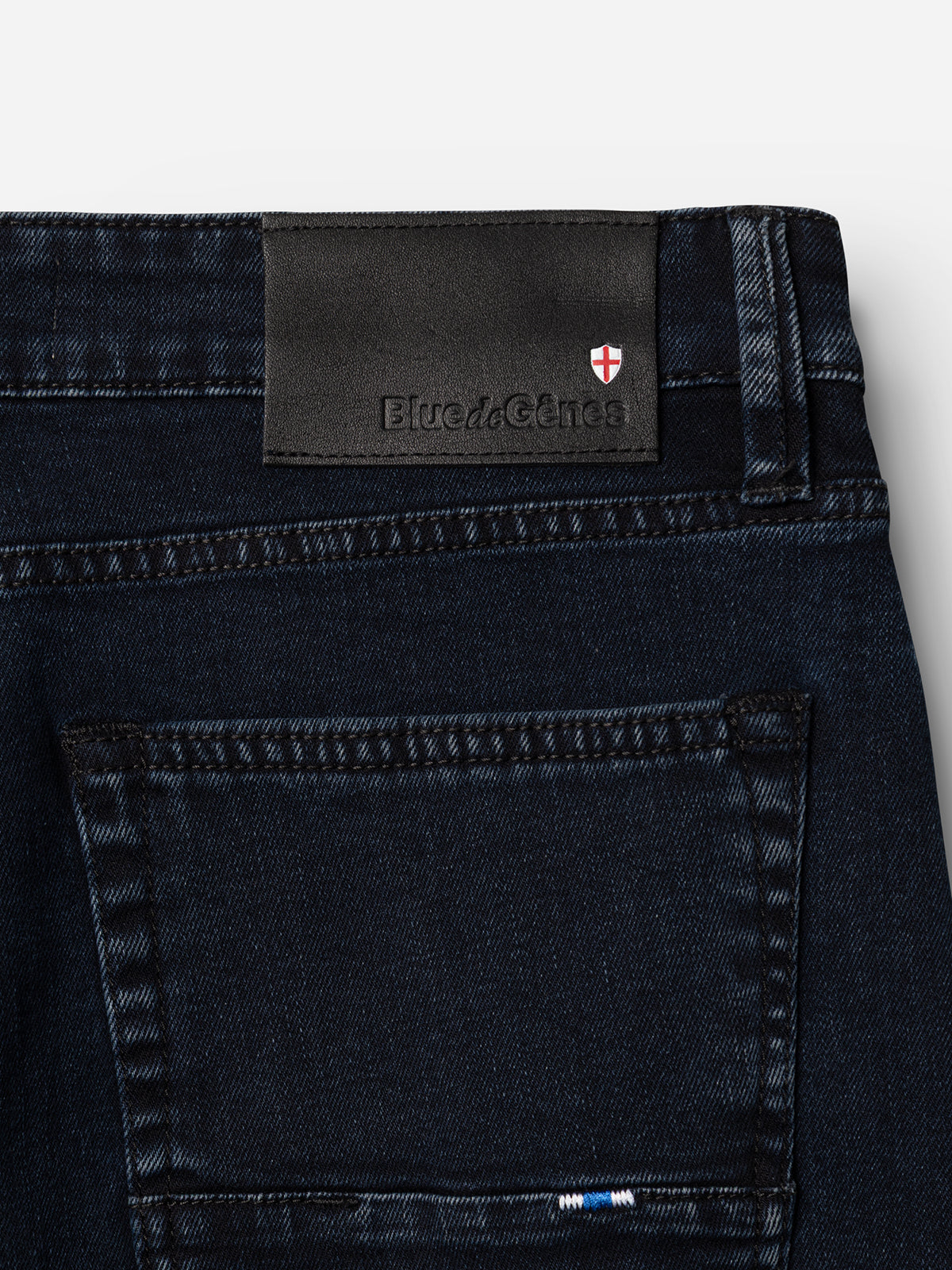 The BGvinci Black OD Jeans by Blue de Gênes feature blue-black stretch denim, a back pocket, and a black leather waistband patch with the Blue de Gênes logo and red and white shield emblem.