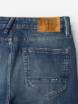 Close-up of the BGvinci Break Jeans - Denim wash by Blue de Gênes, highlighting the comfort stretch denim in dark blue, detailed with visible stitching, a back pocket, a brown leather patch featuring a shield emblem, and part of a belt loop.