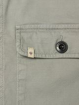 Close-up of the BGpontus Teo Overshirt in Shadow Green by Blue de Gênes, highlighting a textured cotton pocket with a black button engraved with fine text along its edge and a small beige tag adorned with a red shield emblem. This relaxed fit overshirt features subtle detailing for an elevated casual style.