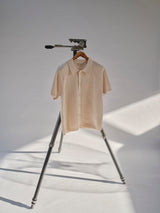 The BGbilly S/S Cardigan - Chalk from Blue de Gênes, a cream-hued short sleeve knitwear shirt with a vintage cardigan-inspired design, hangs from a wooden hanger on a tripod, its texture highlighted by soft light and shadows against a plain backdrop.