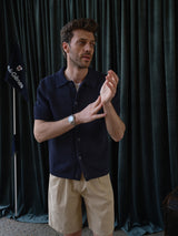 A man wearing the Blue de Gênes BGbilly S/S Cardigan in Dark Navy and beige shorts stands indoors near dark green curtains, adjusting his left sleeve. A black flag with white text is visible in the background.