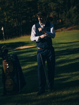 A man wearing the Blue de Gênes BGkendrick Toll Jacket in Navy Stripe stands on a golf course, adjusting his cuff while holding a golf club. A golf bag filled with clubs sits beside him, with trees visible in the background.