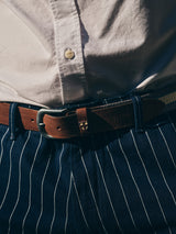 Close-up of a person wearing the Blue de Gênes BGkendrick Toll Jacket in Navy Stripe, paired with a white button-up shirt and a brown leather belt featuring a small crest. Sunlight illuminates the relaxed fit and pinstriped design of the jacket.