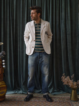 A man stands in front of dark green curtains, wearing a crisp white blazer over the Blue de Gênes BGwally Stripe Polo in Shadow Green, paired with blue jeans and black shoes. A golf bag and a vase with dried flowers are visible nearby.