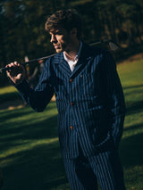 Wearing the Blue de Gênes BGkendrick Toll Jacket in Navy Stripe, a man stands outdoors on the grass, holding a golf club over his shoulder. Sunlight filters through the trees in the background.