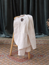 The BGbaio Twill Blazer in Chalk by Blue de Gênes is draped over a wooden chair on a patterned rug, with dark curtains in the background.