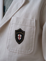 A close-up of the BGbaio Twill Blazer in Chalk by Blue de Gênes, highlighting a white cotton twill pocket adorned with a black and gold shield-shaped patch featuring a red cross on a white background and the Blue de Gênes name.