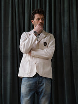 A man wearing the Blue de Gênes BGbaio Twill Blazer in Chalk and jeans stands in front of dark curtains, looking thoughtful with his hand on his chin.