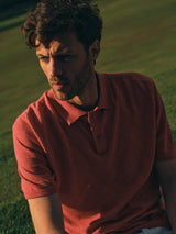 A man with wavy hair wearing the Blue de Gênes BGwally Diamond Polo in Coral sits outdoors on grass in warm, low sunlight, looking down with a thoughtful expression.