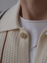 A close-up of a person’s neck wearing a white 100% cotton crew-neck shirt, the Blue de Gênes BGwalter S/S Cardigan in Chalk—a textured cream short sleeve knit cardigan with thin stripes and a button—and a delicate silver chain necklace.