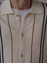 A close-up of a person wearing the BGwalter S/S Cardigan in Chalk by Blue de Gênes—a cream-colored, textured button-up shirt with vertical black and brown stripes, styled over a white T-shirt. Crafted from 100% cotton, the look is finished with a partially visible thin chain necklace.