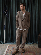 A man wearing the Blue de Gênes BGcricket Lin Blazer in Grey Mel stands on a patterned rug, looking to the side. Dark curtains and a flag on a pole are in the background.