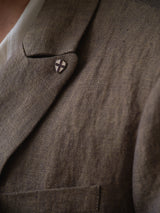 Close-up of the BGcricket Lin Blazer - Grey Mel by Blue de Gênes, a grey double-breasted blazer with a small, shield-shaped pin featuring a cross design on the lapel. A white shirt is partially visible underneath the blazer.