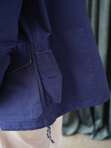 A close-up of the Blue de Gênes BGcanopy Jacket in Navy Peony showcases its relaxed fit, visible stitching, flap pocket, buttonhole, and rope drawstring at the hem. Cream-colored pants and dark green curtains in the background further emphasize the jacket’s utility workwear aesthetic.
