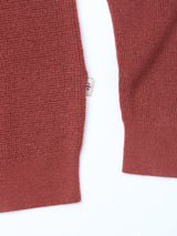 Close-up of the Porto Polo Knit - Henna by Blue de Gênes, a rust-colored textured sweater with ribbed cuffs and hem, shown against a white background. A small fabric tag with a shield emblem appears on the side seam.