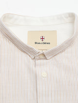 Close-up of the BGbasso Meco Shirt in Beige Stripe, a button-up shirt with beige and white stripes and a stand collar. The Blue de Gênes label features a red and white shield logo on a tan background. Expertly crafted from 100% cotton, this shirt is proudly made in Portugal.