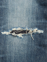 Distressed blue denim from the Giacomo Osaka Jeans – Dark Denim by Blue de Gênes, featuring a horizontal tear that reveals a darker fabric beneath and is bordered by frayed white threads.
