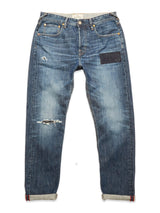 The Giacomo Osaka Jeans by Blue de Gênes in dark denim are a pair of rigid, blue distressed jeans featuring visible rips and a dark patch on the right thigh, shown laid flat with rolled cuffs.