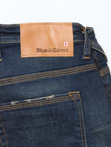 The Repi Dark Vintage Jeans by Blue de Gênes are mid blue comfort stretch denim with yellow stitching, a brown leather patch reading the brand, a red and white shield emblem, regular slim-fit, vintage wash, and a subtly worn back pocket.