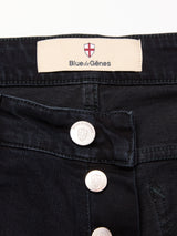 Close-up of Recco BlueBlk Jeans by Blue de Gênes in blue-black denim, showing the waistband label with brand name and red cross emblem. Three silver buttons with the same emblem are visible near the fly, highlighting the relaxed fit.