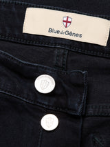Close-up of Recco BlueBlk Jeans by Blue de Gênes in blue-black denim, featuring the brand’s label with a red cross emblem and two metallic buttons engraved with the logo and brand name.