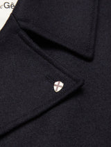 Close-up of the collar of the Shiba Melton Coat in Dark Navy by Blue de Gênes, crafted from luxurious Italian Melton wool. A small, silver, shield-shaped pin with a cross design is attached to the collar, and part of a white label with black text referencing refined Portuguese craftsmanship can be seen in the background.