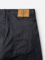 Close-up of the back pocket of Blue de Gênes BGleroy Jeans - Raw Denim, showcasing a tan leather patch with a shield emblem stitched above the pocket against a white background. The image highlights the innovative raw denim construction and the unique absence of an outer leg seam, emphasizing both style and craftsmanship.
