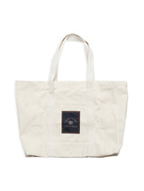 The BDG Canvas Bag - Off White by Blue de Gênes is a spacious tote with two handles and a square dark front patch featuring text and a small emblem.
