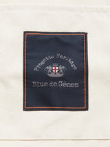A dark blue fabric patch with "Progetto Heritage Blue de Gênes" and a crest is sewn onto the off-white BDG Canvas Bag by Blue de Gênes, featuring orange stitching.