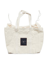 The Blue de Gênes BDG Canvas Bag - Off White is a cream-colored tote with handles and a drawstring, featuring a black rectangular patch with white text and a small graphic on the front. The bag is shown on a white background.
