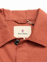 A close-up of the BGpontus Teo Overshirt in Copper Brown by Blue de Gênes showcases its rust-hued, textured cotton design with a collared neckline and black button. The overshirt features a small red and white shield logo above the brand name tag, emphasizing its relaxed fit and refined detailing.