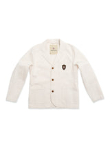 The BGbaio Twill Blazer - Chalk by Blue de Gênes is a white, classic cotton blazer featuring brown buttons, two front pockets, and a black-and-red shield-shaped patch on the left chest. This regular fit blazer is shown laid flat against a white background.