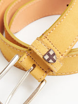 A close-up of the Blue de Gênes Suede Belt - Yellow Grain shows light yellow suede, beige stitching, a silver buckle, and a small metallic emblem with a white cross on a dark background.