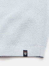 Close-up of the Blue de Gênes Filo Knit Polo - Blue Water, showing its light blue textured fabric and a small navy hem tag with an orange crown and white shield motif.