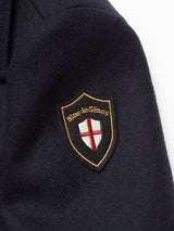 The Peacoat - Dark Navy by Blue de Gênes features a navy blue fabric with a shield-shaped patch that reads "Blue de Gênes" above a red cross on a white background, finished with gold stitching.