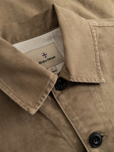 A close-up of the BGcuddy Nebraska Jacket in brown by Blue de Gênes, featuring a tan high-density cotton fabric, black buttons, and a collared design. The inside label displays the Blue de Gênes name with a small red and white shield logo above the text.