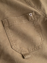 Close-up of a brown high-density cotton fabric pocket with detailed stitching on the Blue de Gênes BGcuddy Nebraska Jacket. A small pin, showcasing a white shield emblazoned with a black cross, is attached near the pocket’s top right corner—an accent that underscores the jacket’s thoughtful blend of functional style and craftsmanship.