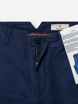 Close-up of the BGliam Nebraska Chino - Blue by Blue de Gênes, crafted from navy blue cotton with a high-density dobby weave that adds subtle texture. The trousers feature a button and zipper fly, shown partially open to reveal the inner label and a piece of paper tucked inside the waistband. An adjustable buckle strap at the waist ensures a comfortable fit.