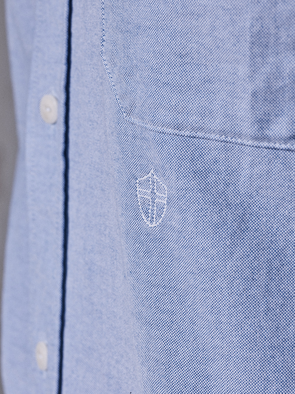Close-up of the BGsandro Oxford Shirt - Blue by Blue de Gênes, featuring white buttons and a white embroidered shield logo on the chest pocket.