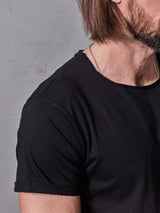 A close-up of a man's upper body, wearing the Blue de Gênes Sagi Nuovo T-shirt in black—a true wardrobe staple—and a silver chain necklace, with brown hair and a beard. Only his right arm, shoulder, and part of his face are visible against a plain background.