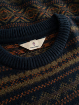 A close-up of the BGrhys Knit - Dark Navy sweater collar reveals a vintage-inspired woven pattern in blue, brown, and orange tones. Inside the crewneck, a tag displays Blue de Gênes along with a small shield logo above the text.