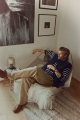 A man with a beard relaxes in a modern chair with a fur cover, surrounded by abstract black-and-white artwork on the wall. He wears a striped shirt, vest, Blue de Gênes BGliam Nebraska Chino - Brown trousers with an adjustable buckle strap, and boots in a cozy, stylish room.